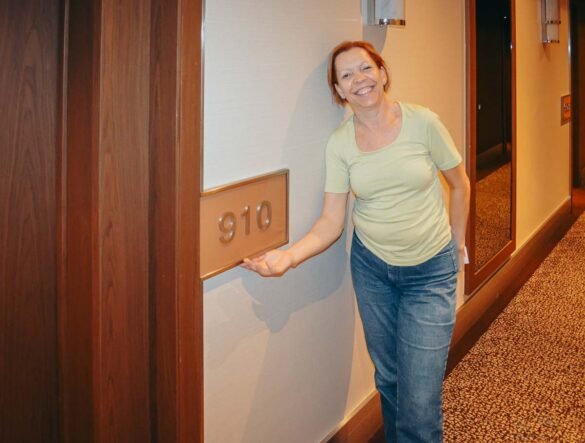 Mulher de camiseta verde sorri ao lado da placa do quarto 910 em um corredor de hotel.