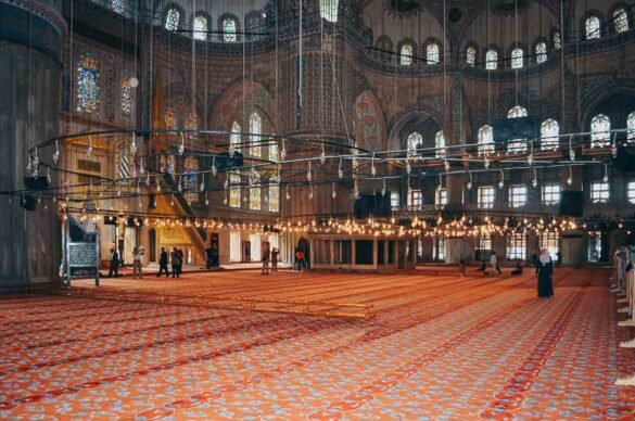 Visão ampla do interior da Mesquita Azul com tapetes vermelhos estampados e vitrais coloridos.
