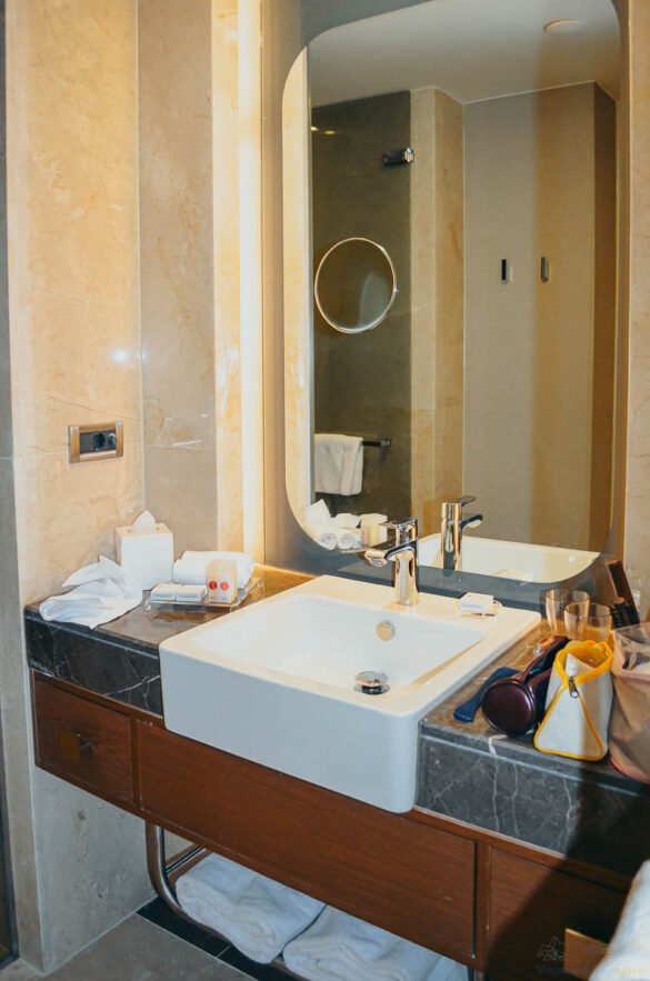 Bancada de mármore com pia branca, espelho iluminado e itens de higiene organizados.