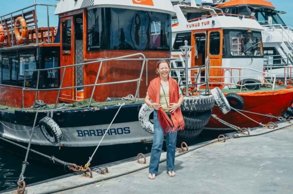 Mulher em frente ao barco de madeira chamado Barbaros em um porto ensolarado.