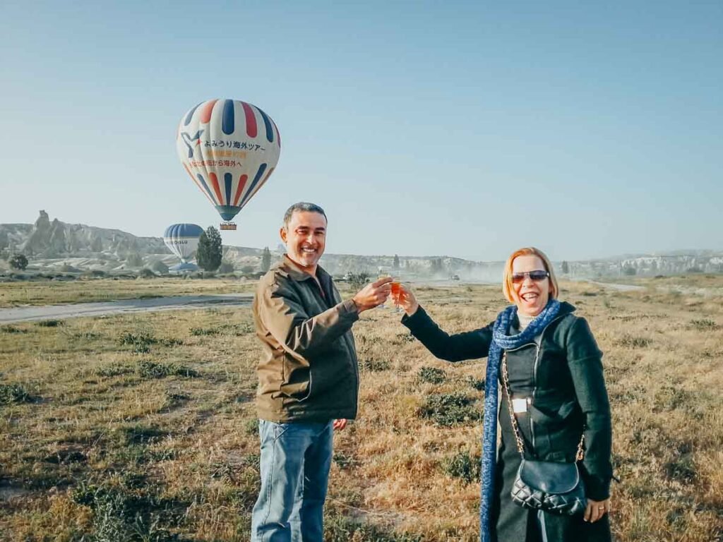 Casal brinda com taças em campo aberto com balões de ar quente ao fundo na Capadócia.