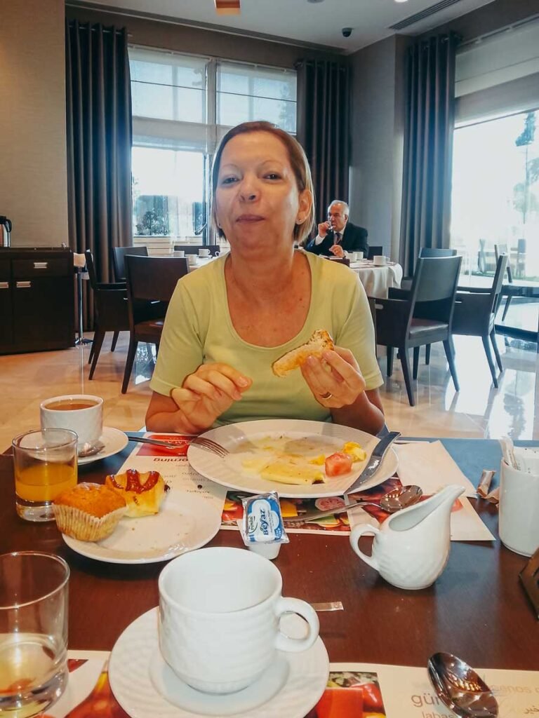 Mulher tomando café da manhã em hotel na Turquia, segurando um pão, com café e suco na mesa.