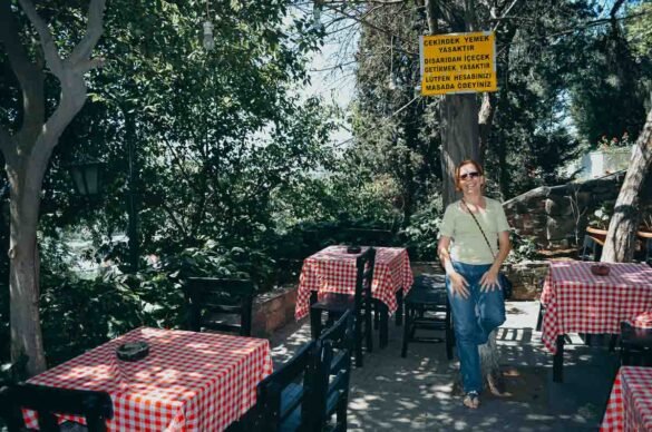 Mulher sorrindo em café com toalhas xadrez vermelhas e placas em turco ao fundo.