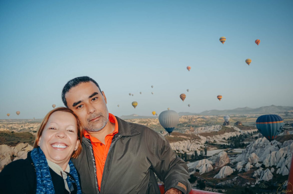 Selfie de casal sorrindo dentro de um cesto de balão com o vale da Capadócia ao fundo.