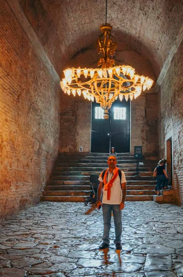 Homem sob grande lustre ornamentado em corredor de pedra com teto em abóbada.