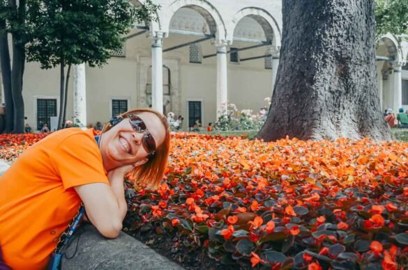 Mulher deitada perto de um mar de flores vermelhas com arcos de um palácio ou mesquita ao fundo.