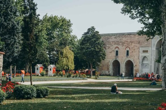 Jardim arborizado com flores vermelhas e arcos de pedra do Palácio Topkapi ao fundo.