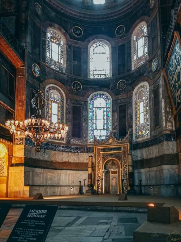 Altar Mihrab dourado cercado por vitrais coloridos e grandes lustres circulares.