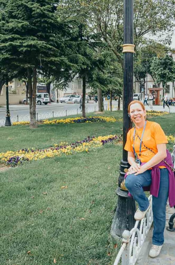 Mulher de camiseta laranja sorrindo em um jardim florido no Parque Sultanahmet.