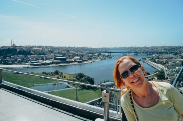 Mulher ruiva sorrindo em close com a vista aérea do Chifre de Ouro ao fundo sob céu azul.