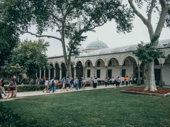 Pátio arborizado com arcos e cúpulas brancas ao fundo, com turistas caminhando em Istambul.