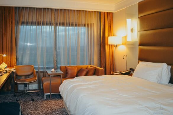 Quarto de hotel moderno com cama branca, poltrona de couro e iluminação quente em tons de âmbar.