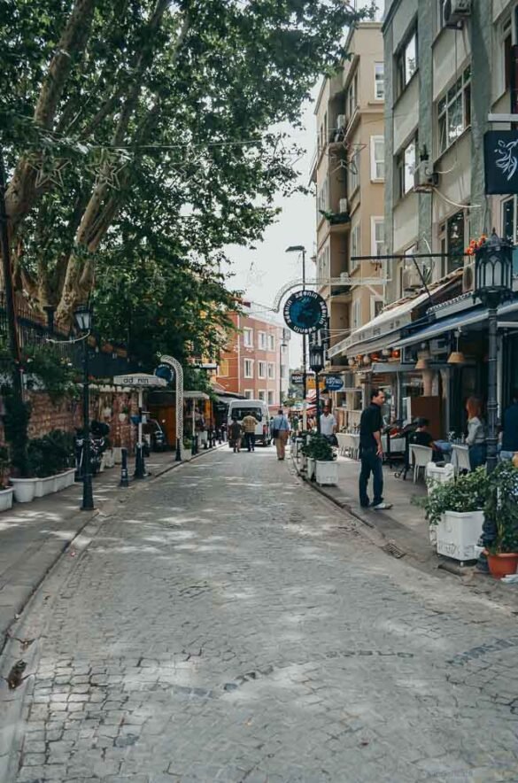 Rua estreita de paralelepípedos com árvores altas, prédios baixos e mesas de café na calçada.