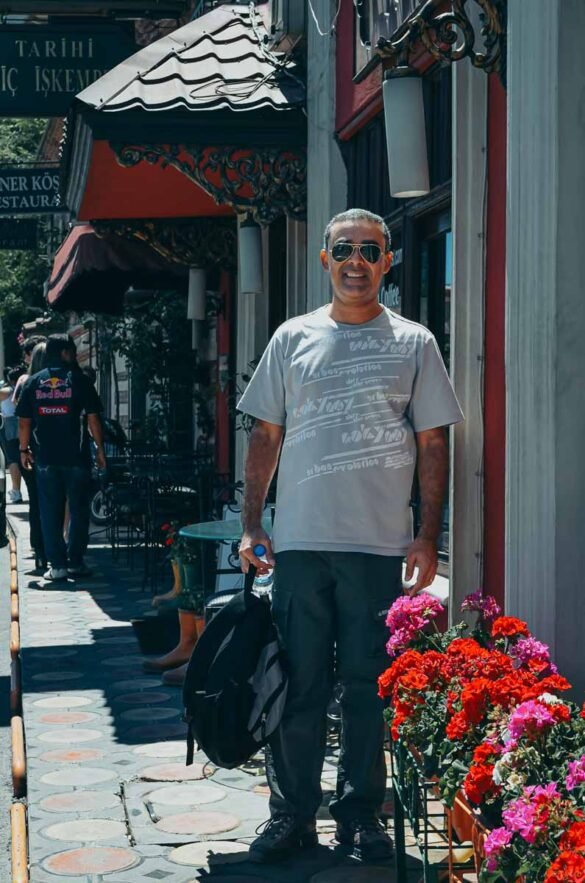 Homem em pé em calçada de rua turca com flores vermelhas e arquitetura local ao fundo.