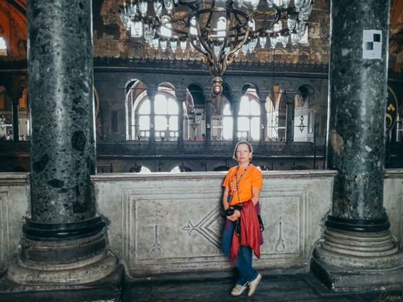 Mulher posando na galeria superior da Hagia Sophia, com lustres e arcos ao fundo.