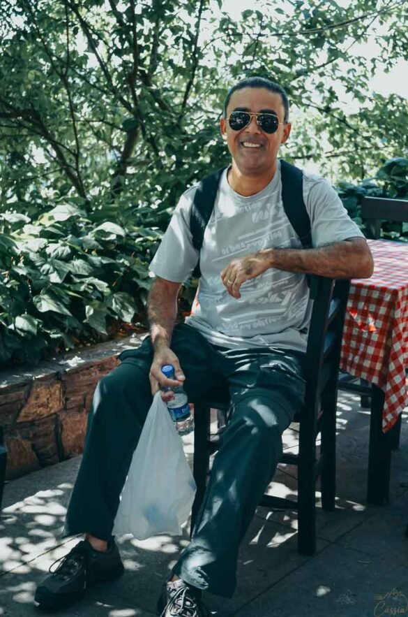 Homem de óculos escuros sentado em mesa de café ao ar livre sob a sombra das árvores.