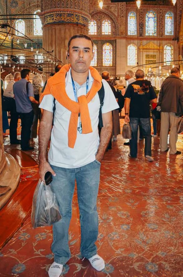 Homem com suéter laranja nos ombros posa no tapete decorado da Mesquita Azul em Istambul.