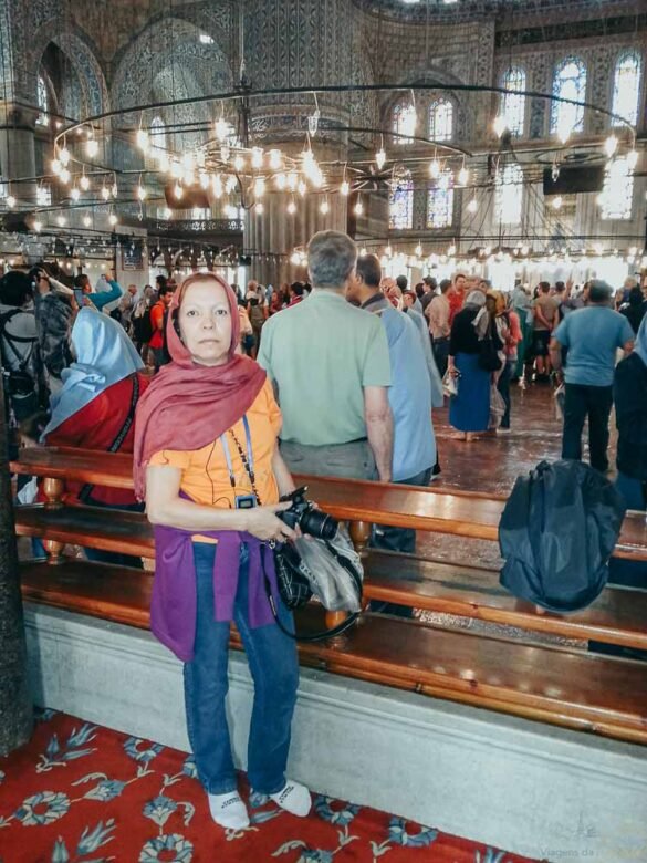 Mulher com lenço vermelho posa dentro da Mesquita Azul, sob lustres circulares e teto decorado.