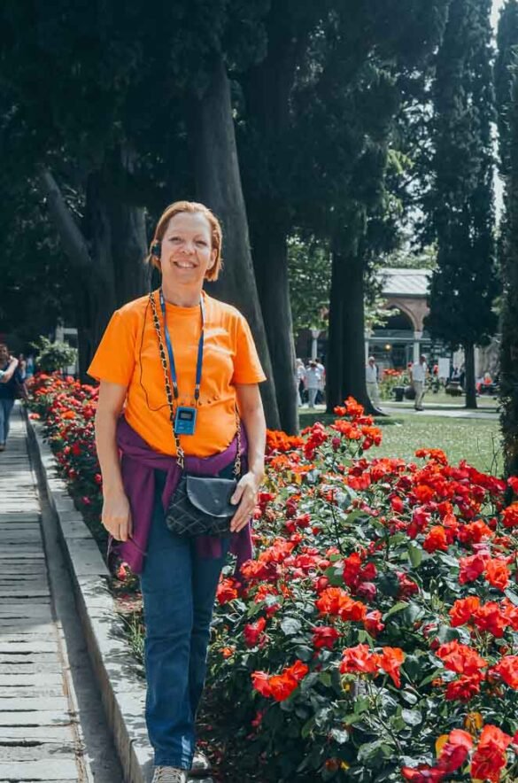 Mulher de blusa laranja sorrindo ao lado de um longo canteiro de rosas vermelhas em um jardim.
