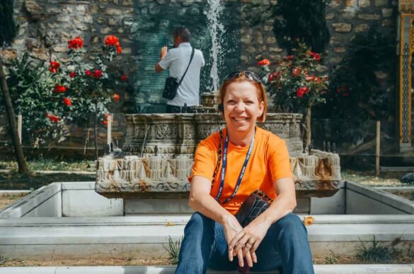 Mulher sentada em degraus de pedra à frente de um chafariz ornamental antigo esculpido.