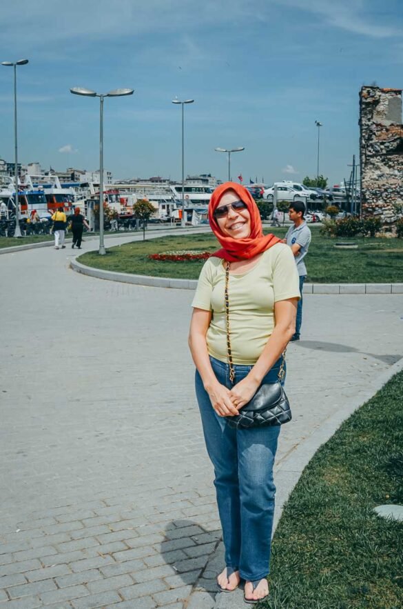 Mulher sorridente com lenço vermelho e óculos escuros em área portuária de Istambul.