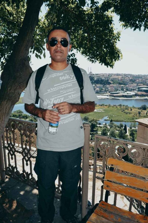 Homem em pé sob sombra de árvore em terraço com parapeito decorado e vista para cidade.