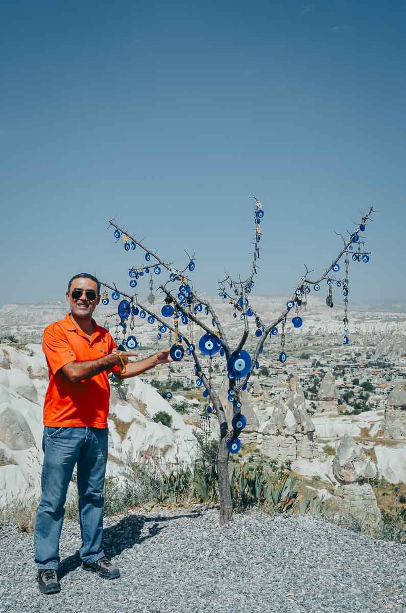 arvore-olho-grego-nazar-cappadocia-turquia Homem apontando para árvore com amuletos de olho grego na Capadócia.