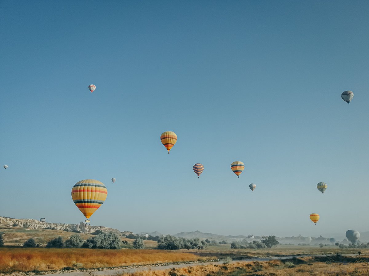 baloes-ceu-azul-campo-dourado-capadocia Vários balões de ar quente flutuando sob um céu azul limpo acima de um campo dourado.