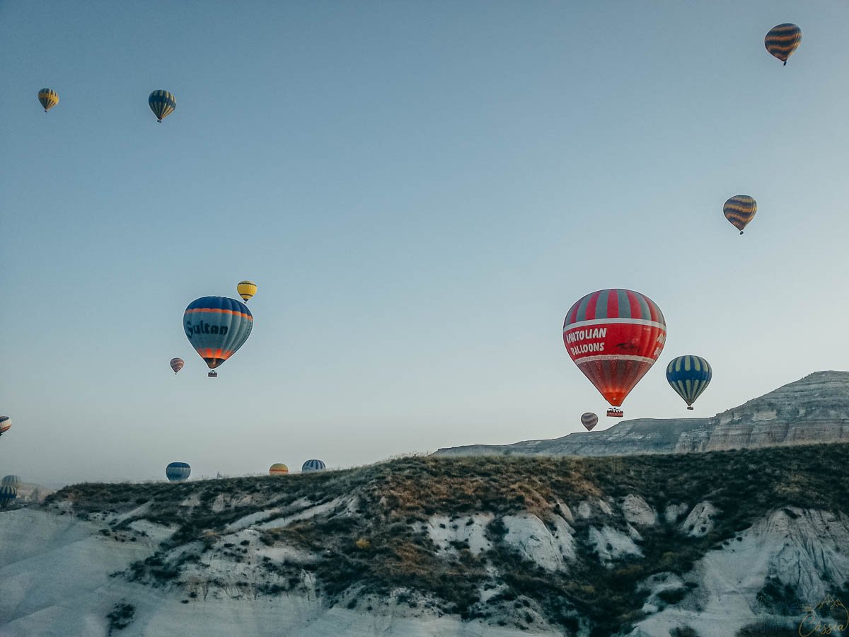baloes-coloridos-ceu-azul-capadocia Balões de ar quente de diversas cores voando próximos a um cume rochoso sob um céu azul claro.