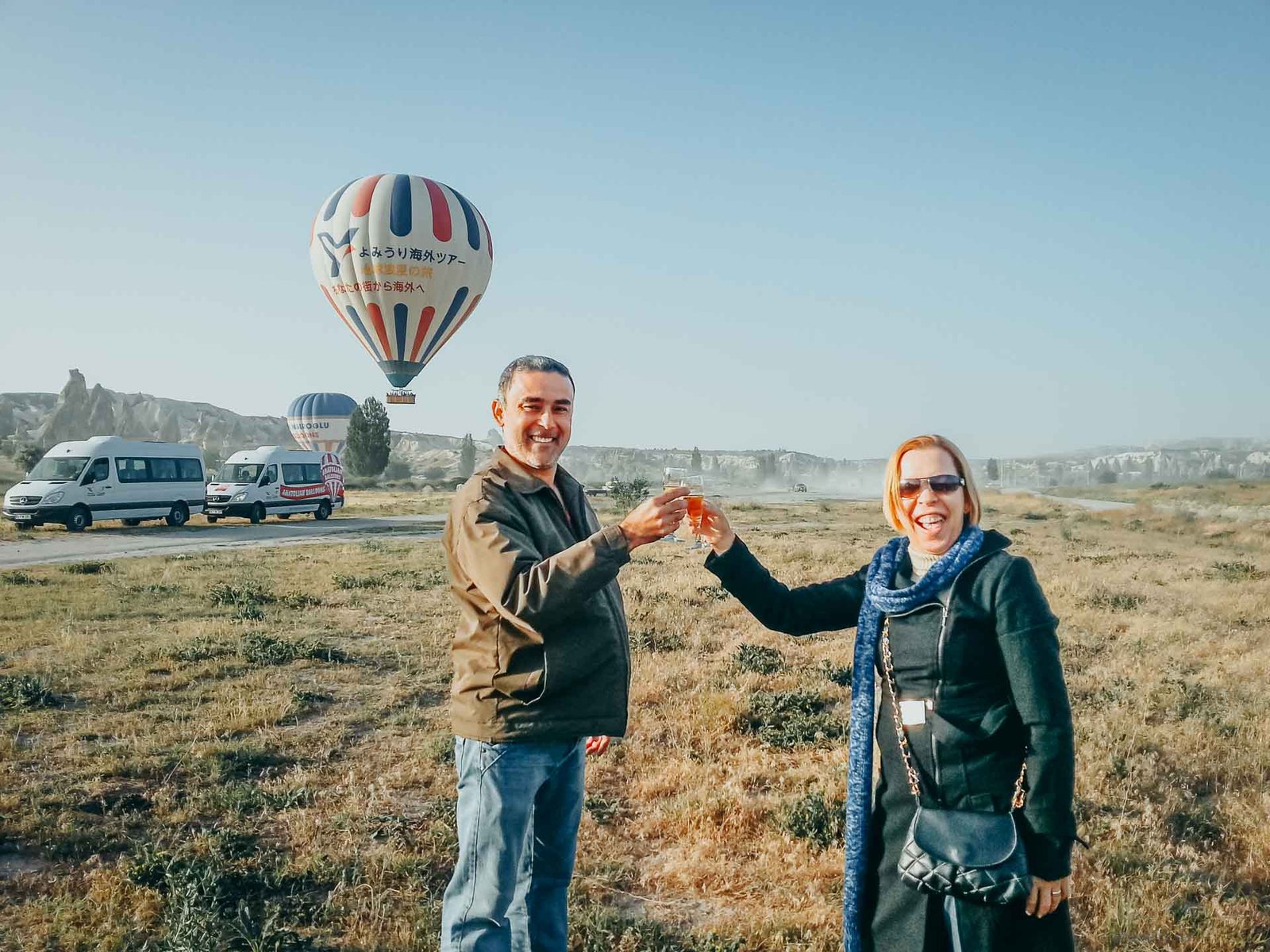 brinde-pouso-balao-capadocia Homem e mulher sorrindo e brindando com taças num campo após o pouso do balão.