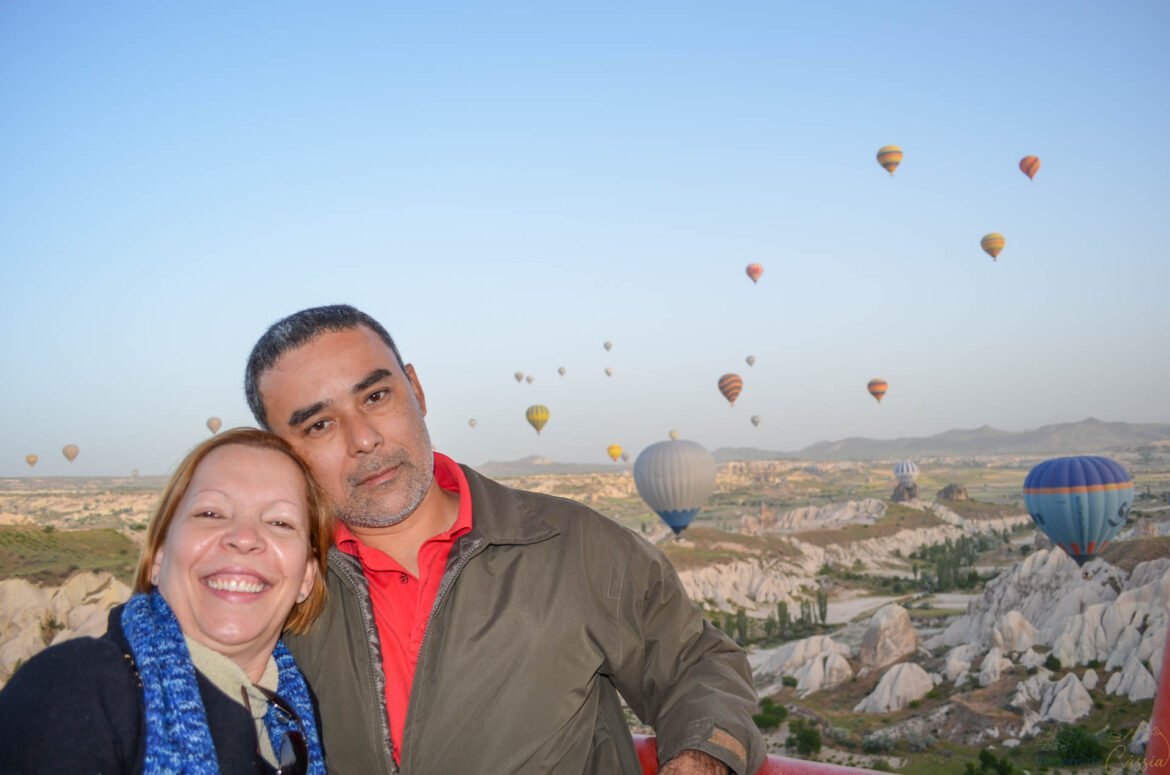 Casal sorrindo em selfie com vários balões de ar quente no céu da Capadócia ao fundo.