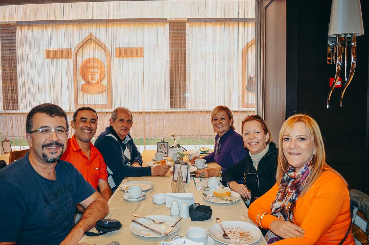 grupo-amigos-mesa-cafe-restaurante Seis pessoas sorrindo enquanto tomam café da manhã juntos em uma mesa de restaurante.