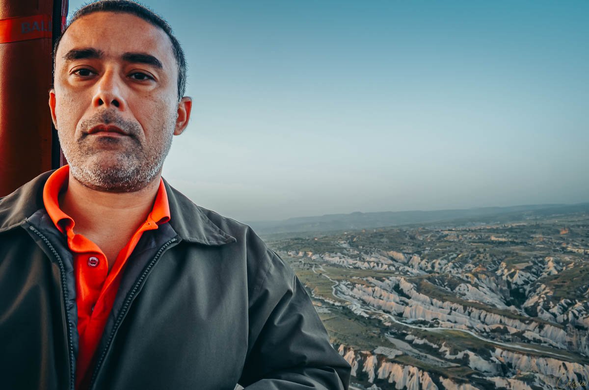 homem-cesta-balao-vista-capadocia Homem com jaqueta cinza e camisa laranja posando sério dentro da cesta de um balão, com o vasto vale da Capadócia ao fundo.