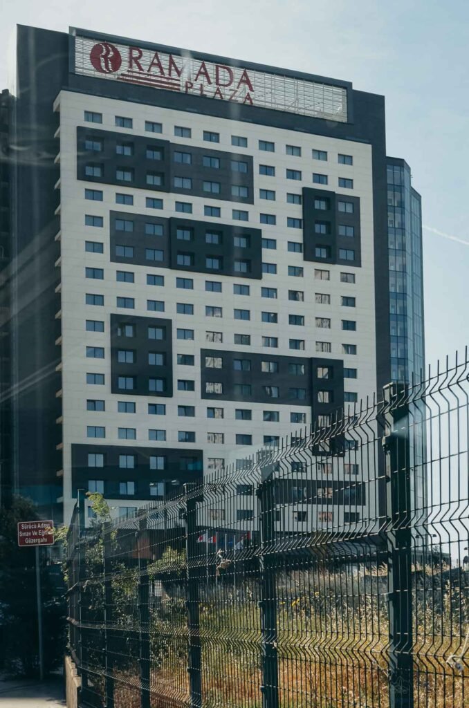 Fachada moderna do hotel Ramada sob céu limpo, vista através de um cercado.