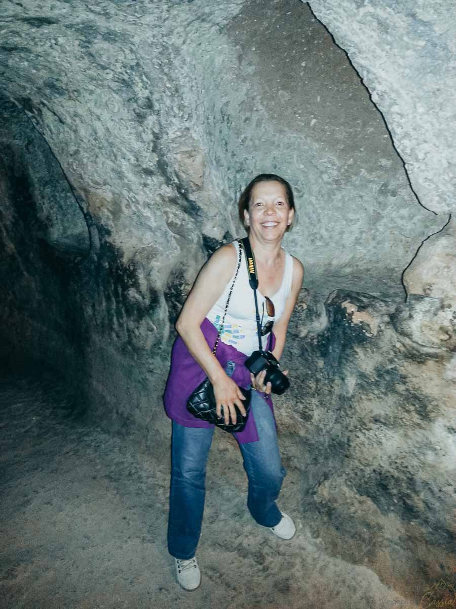 mulher-explorando-caverna-cidade-subterranea Mulher sorrindo com câmera no pescoço explorando o interior de uma caverna de pedra escavada.