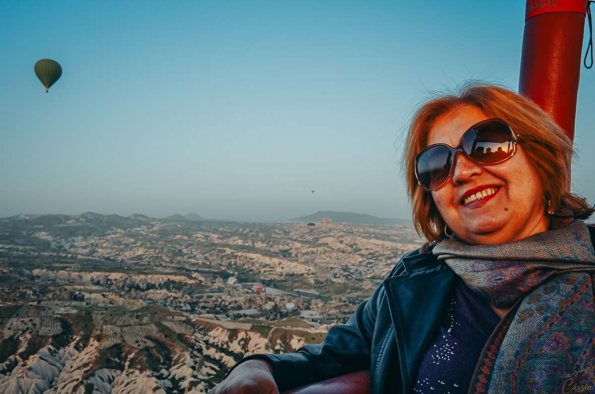 mulher-sorrindo-voo-balao-capadocia Mulher sorridente com óculos escuros e lenço dentro da cesta de um balão, com as formações rochosas da Capadócia ao fundo.