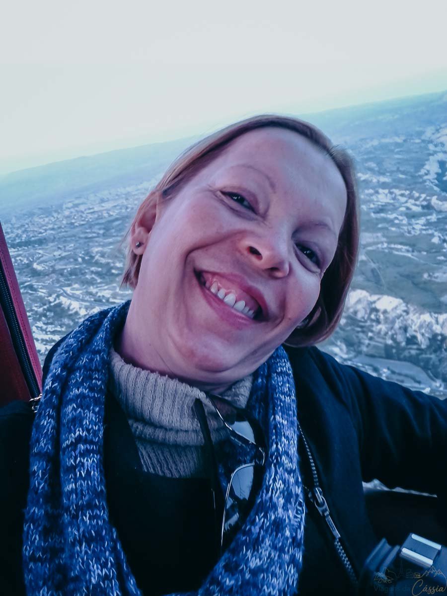 mulher-sorrindo-voo-balao-capadocia Mulher loira sorrindo com cachecol azul tirando foto dentro da cesta de um balão em voo.