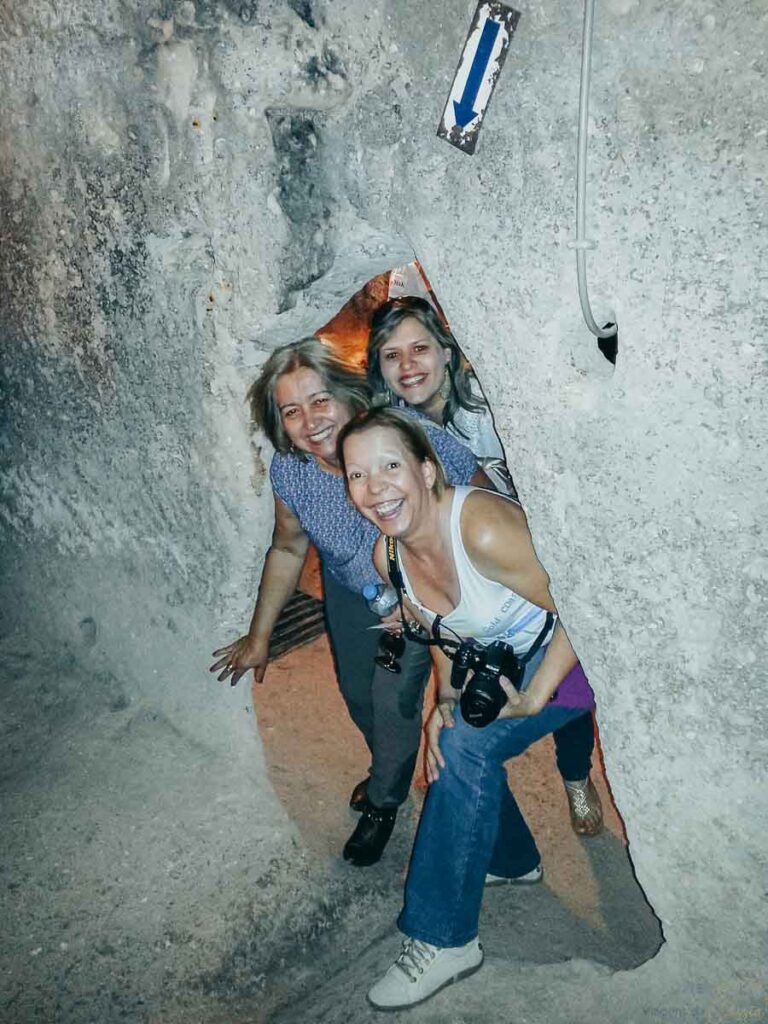Três mulheres sorrindo ao cruzar uma passagem estreita de pedra escavada.