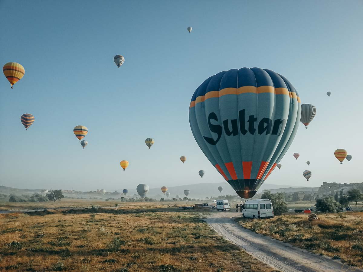 pouso-baloes-campo-van-apoio-capadocia Balões de ar quente descendo em um campo aberto de vegetação rasteira, onde vans e equipes de apoio aguardam em estrada de terra.
