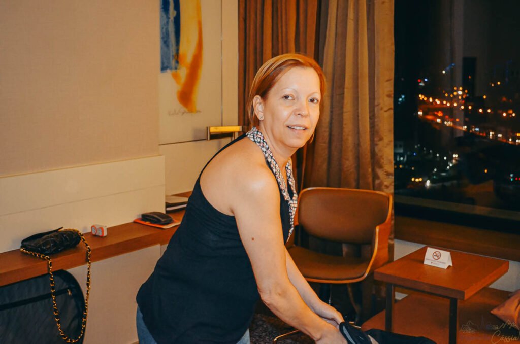 Mulher organizando pertences no quarto de hotel com vista noturna da cidade iluminada ao fundo.