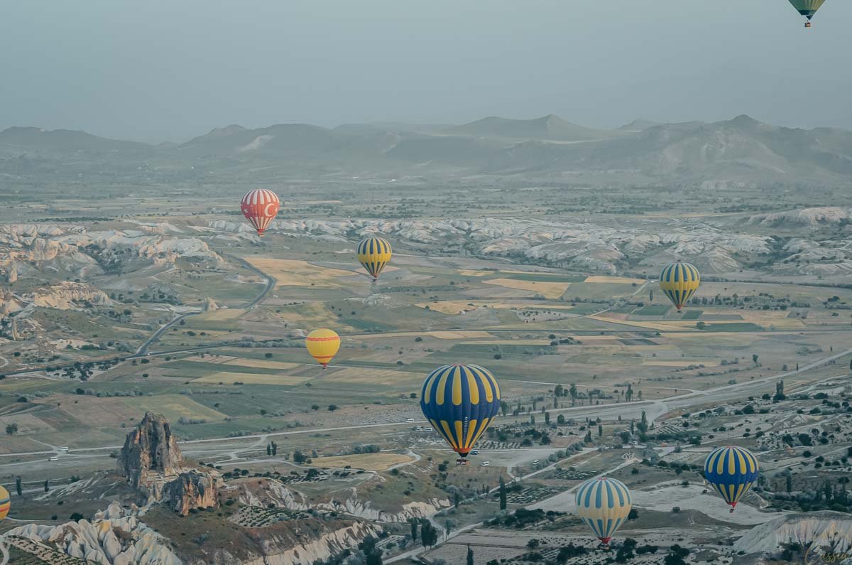 vista-panoramica-baloes-vale-capadocia Vista panorâmica de dezenas de balões espalhados por um vasto vale verde e rochoso na Capadócia durante a névoa matinal.