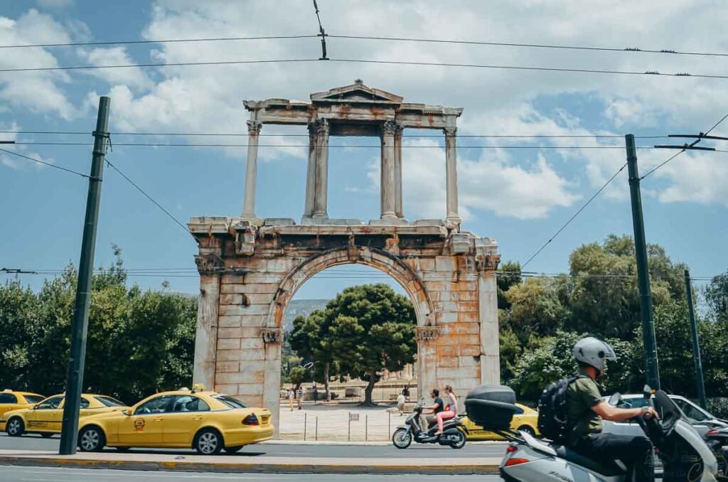 Arco de Adriano em pedra antiga à beira de rua movimentada com táxis amarelos e motos.