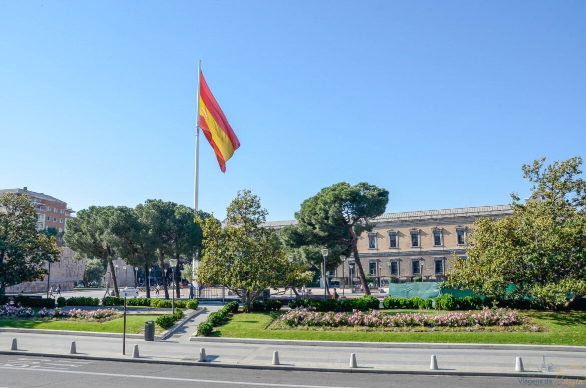 Bandeira da Espanha hasteada em praça arborizada com edifícios ao fundo sob céu azul.