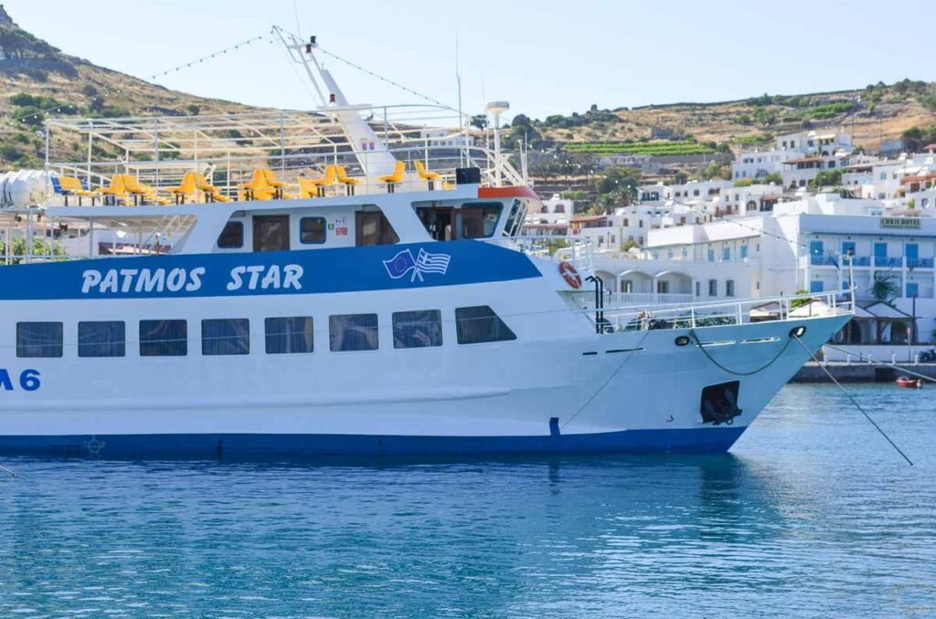 Barco de passageiros Patmos Star atracado em águas azuis com casario branco ao fundo.