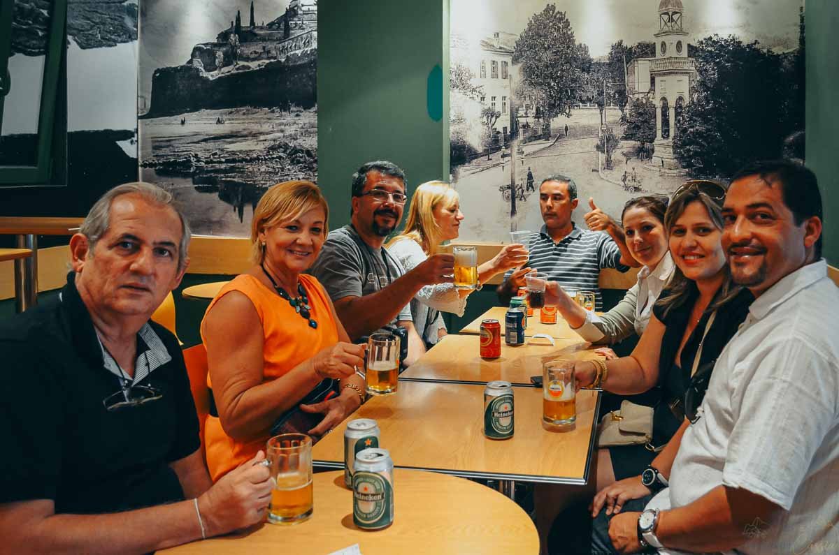 brinde-grupo-viajantes-restaurante-atenas Grupo de amigos reunido ao redor da mesa brindando com copos e latas de cerveja em restaurante.
