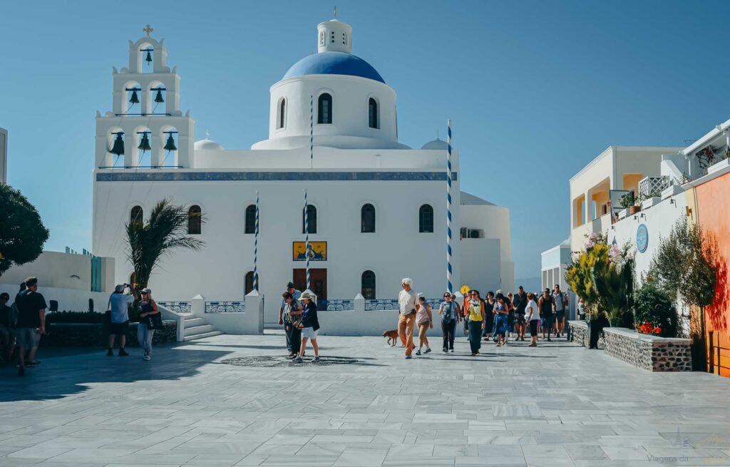 Catedral branca com cúpula azul e campanário com sinos em praça de pedra com pedestres.