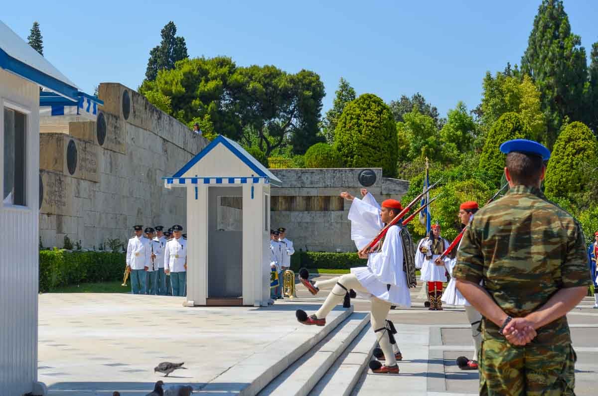 cerimonia-troca-da-guarda-evzones-atenas Guardas Evzones executando o passo cerimonial sincronizado enquanto oficial em farda observa.