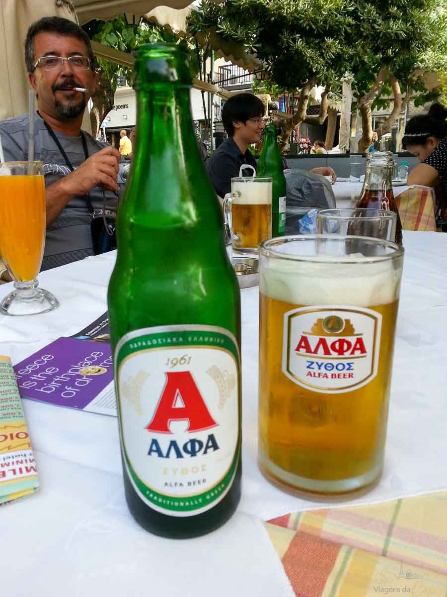 cerveja-alfa-greca-restaurante-atenas Garrafa e copo de cerveja grega Alfa com espuma na mesa de restaurante ao ar livre.