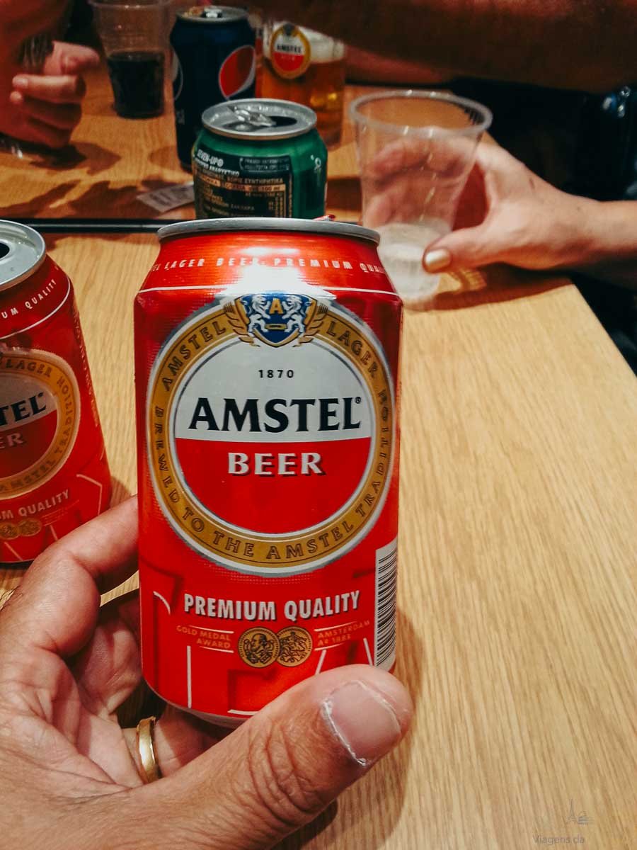 cerveja-amstel-lata-mesa-grecia Mão segurando lata vermelha de cerveja Amstel sobre mesa de madeira em confraternização.
