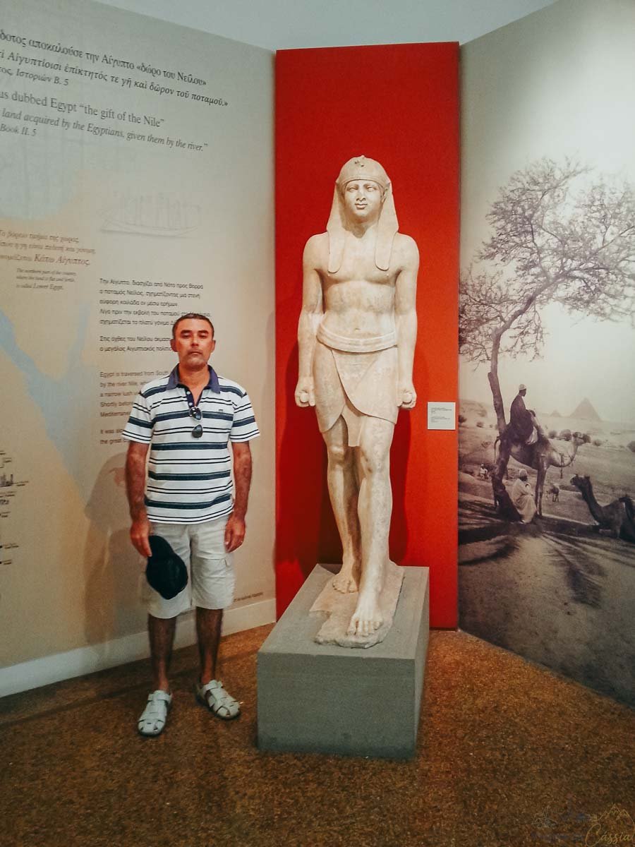 colecao-egipcia-museu-arqueologico-nacional-atenas Homem posando ao lado de estátua de faraó egípcio em exposição no museu de Atenas.
